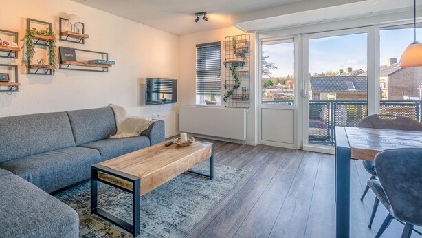 A modern living room with a grey sofa, wooden coffee table, and a dining area with a table and chairs. There are shelves with decorative items and a view through large windows. - Nice Apartment Near the Center of De Koog (De Koog)