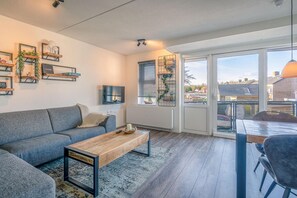A modern living room with a grey sofa, wooden coffee table, and a dining area with a table and chairs. There are shelves with decorative items and a view through large windows. - Nice Apartment Near the Center of De Koog (De Koog)