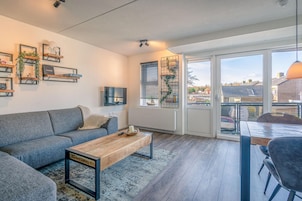 Ein modernes Wohnzimmer mit grauem Sofa, Holztisch und Essbereich mit Tisch und Stühlen. Regale mit Dekorationselementen und ein Blick durch große Fenster.