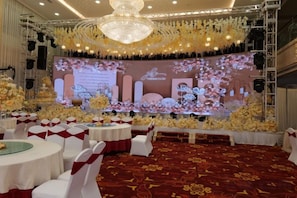 Un salón de banquetes con mesas redondas, sillas blancas y un escenario con decoración floral.