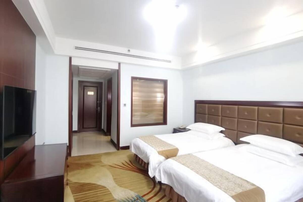 Une chambre d’hôtel avec deux lits, une télévision, une fenêtre avec des stores et un sol recouvert de moquette.