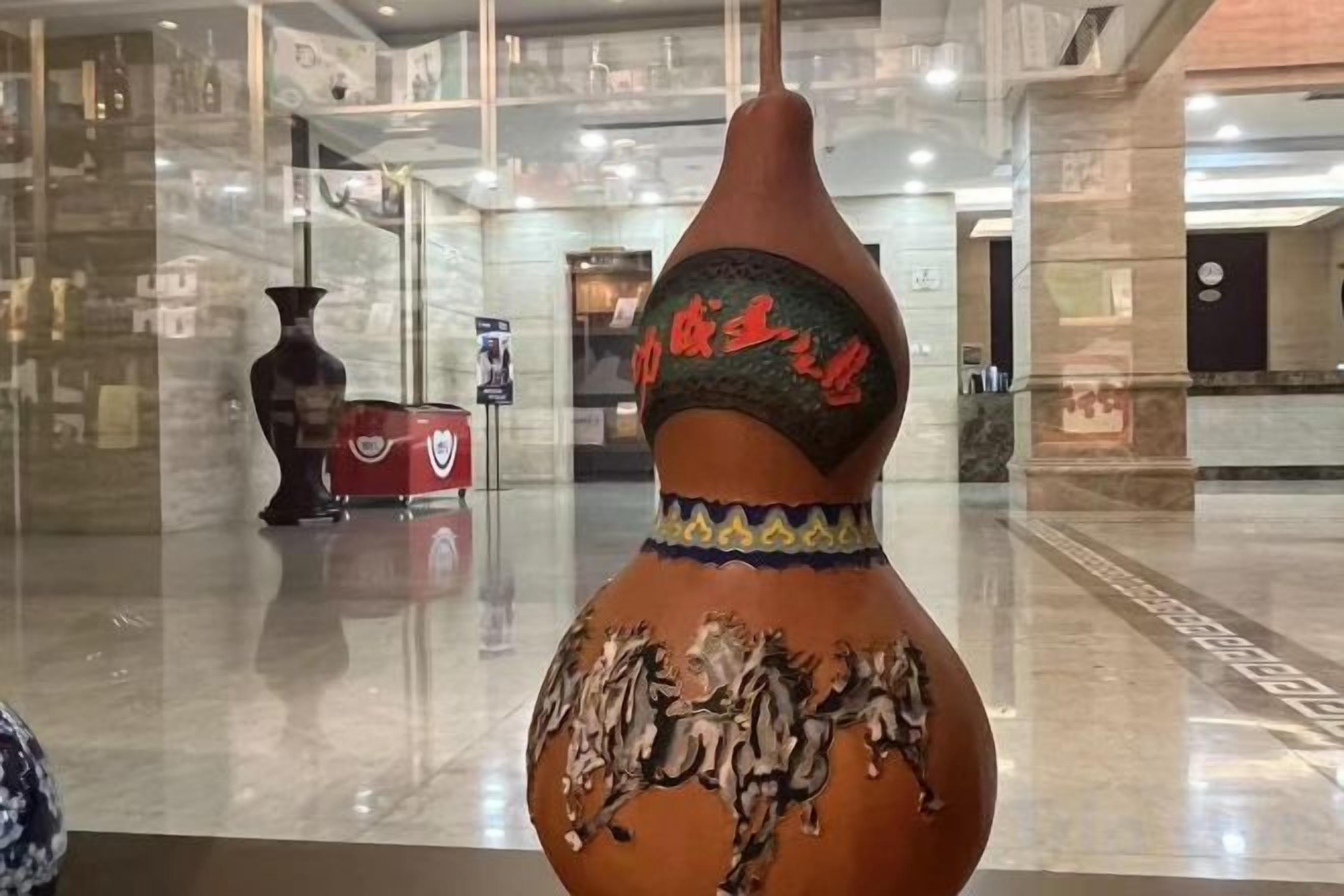 Um vaso grande e ricamente decorado em um saguão com piso lustroso e um vaso vermelho ao fundo.