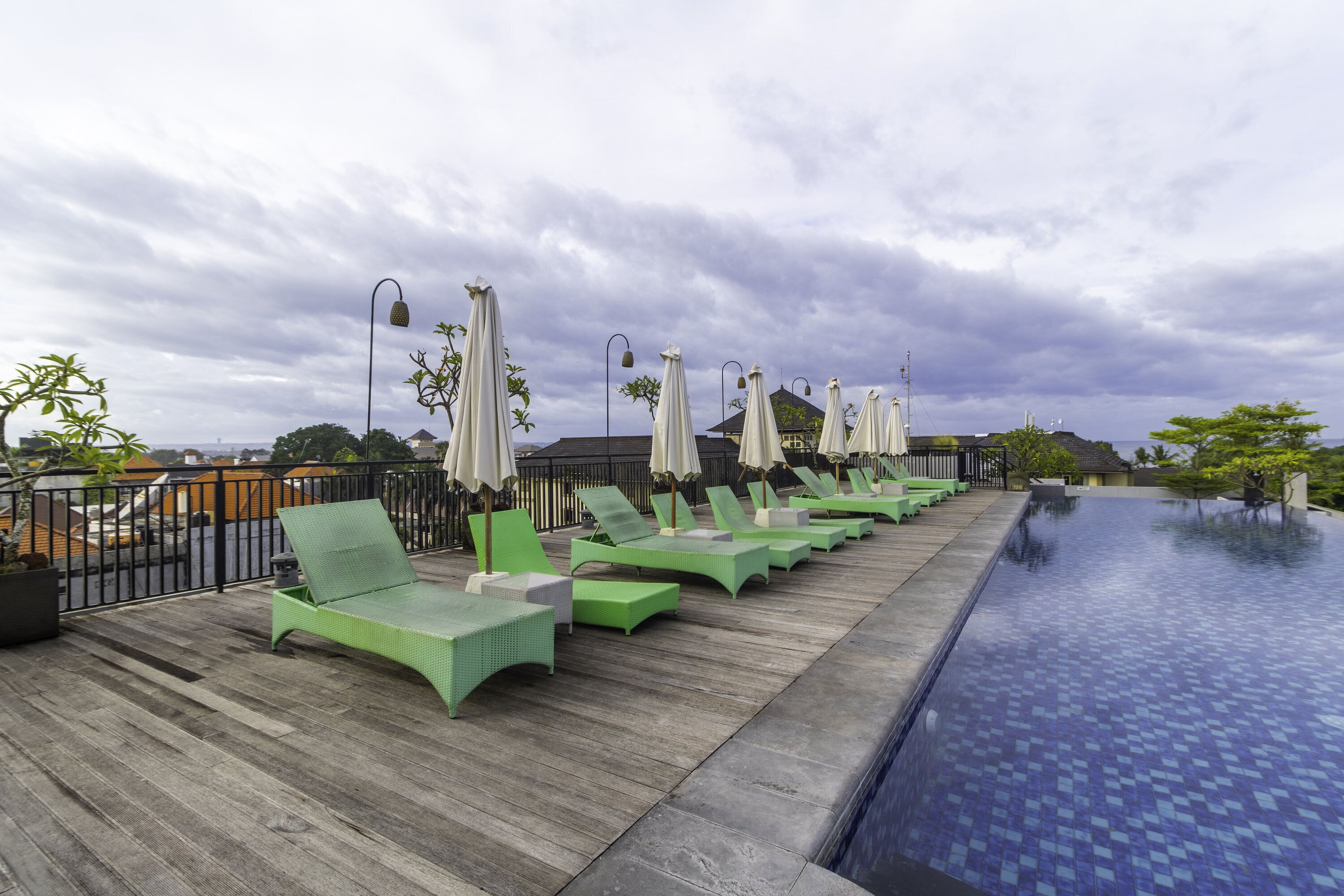 A rooftop pool area with green lounge chairs and umbrellas, surrounded by a wooden deck and a metal railing.