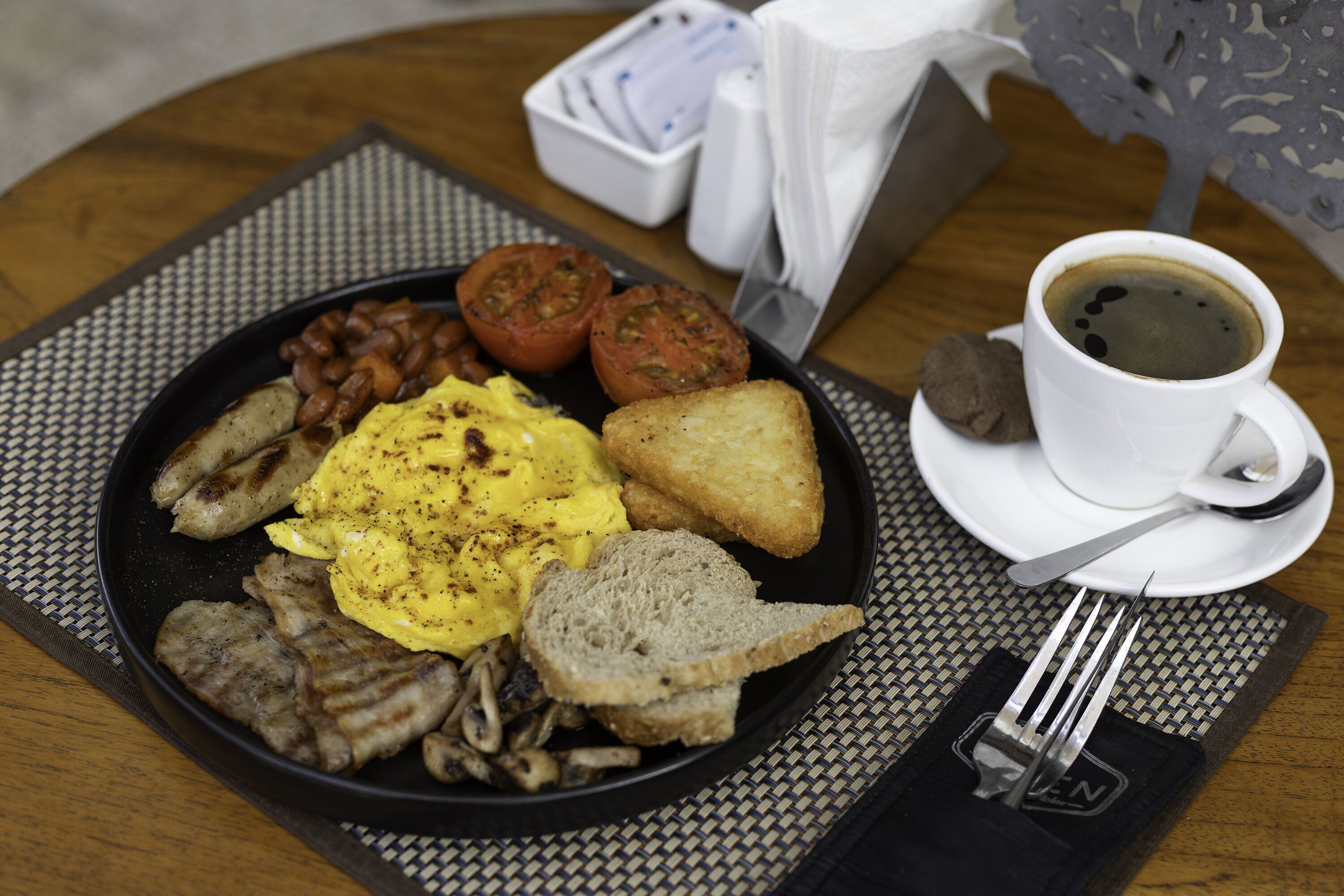 A breakfast plate with eggs, bacon, beans, tomatoes, toast, and a cup of coffee on a table.
