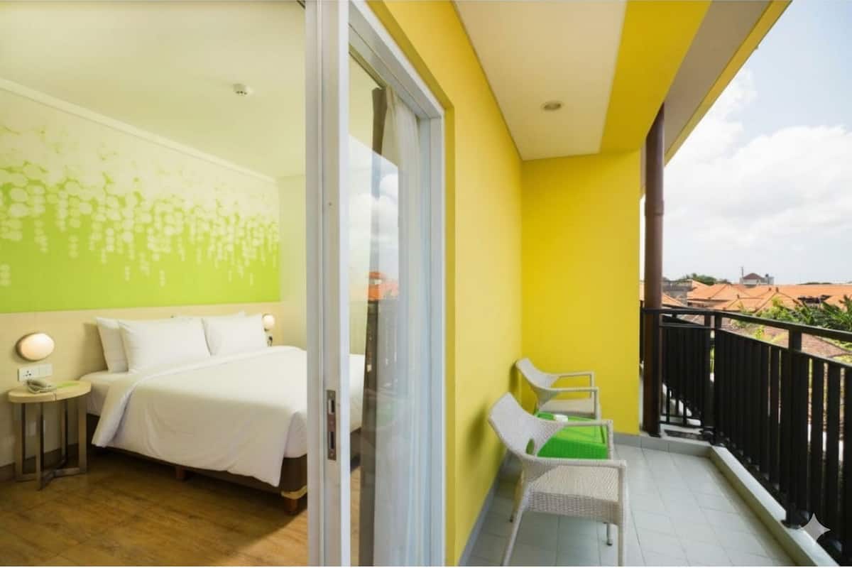A hotel room with a bed, a small table with a lamp, a balcony with a chair, and a view of the city.