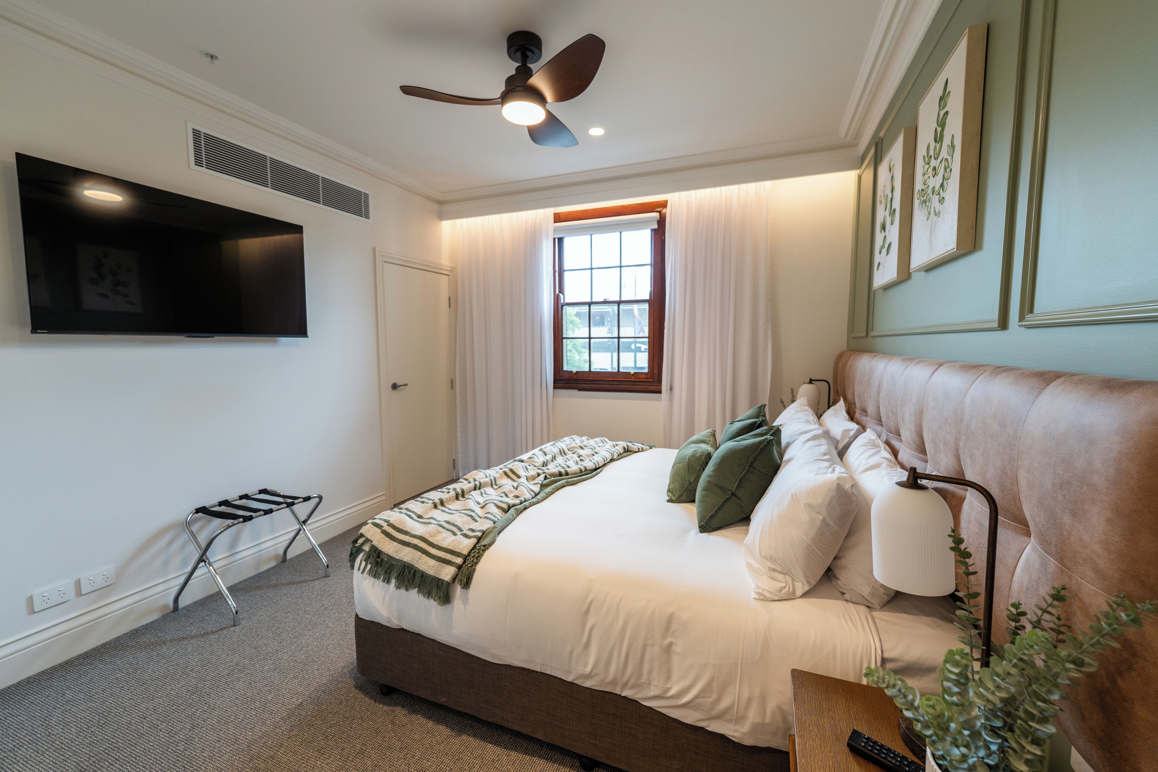 Un dormitorio con cama, ventilador de techo, ventana con cortinas, televisor montado y mesita de noche con control remoto.