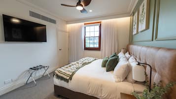 Un dormitorio con cama, ventilador de techo, ventana con cortinas, televisor montado y mesita de noche con control remoto.