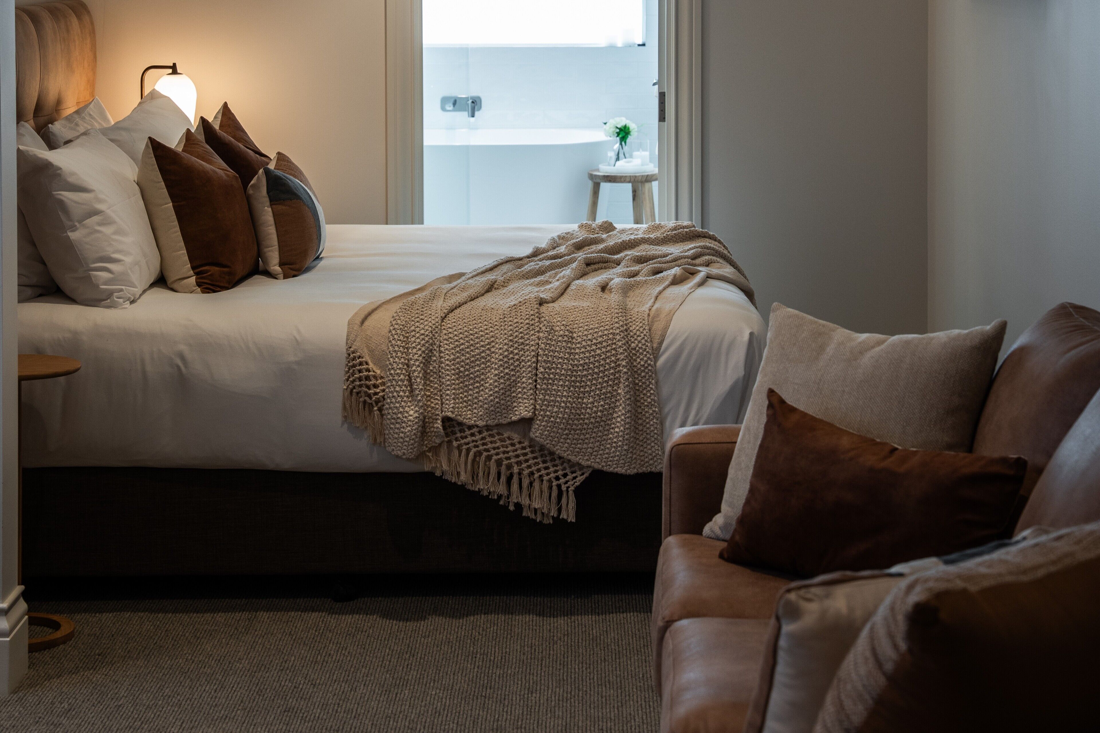 Un dormitorio con cama, mesita de noche, lámpara, ventana y vistas al baño, además de un sofá de cuero marrón.