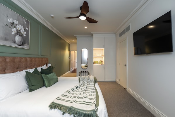 A bedroom with a bed, green pillows, a ceiling fan, a TV, and a hallway leading to a bathroom. - Camden Hotel Suites (Camden)