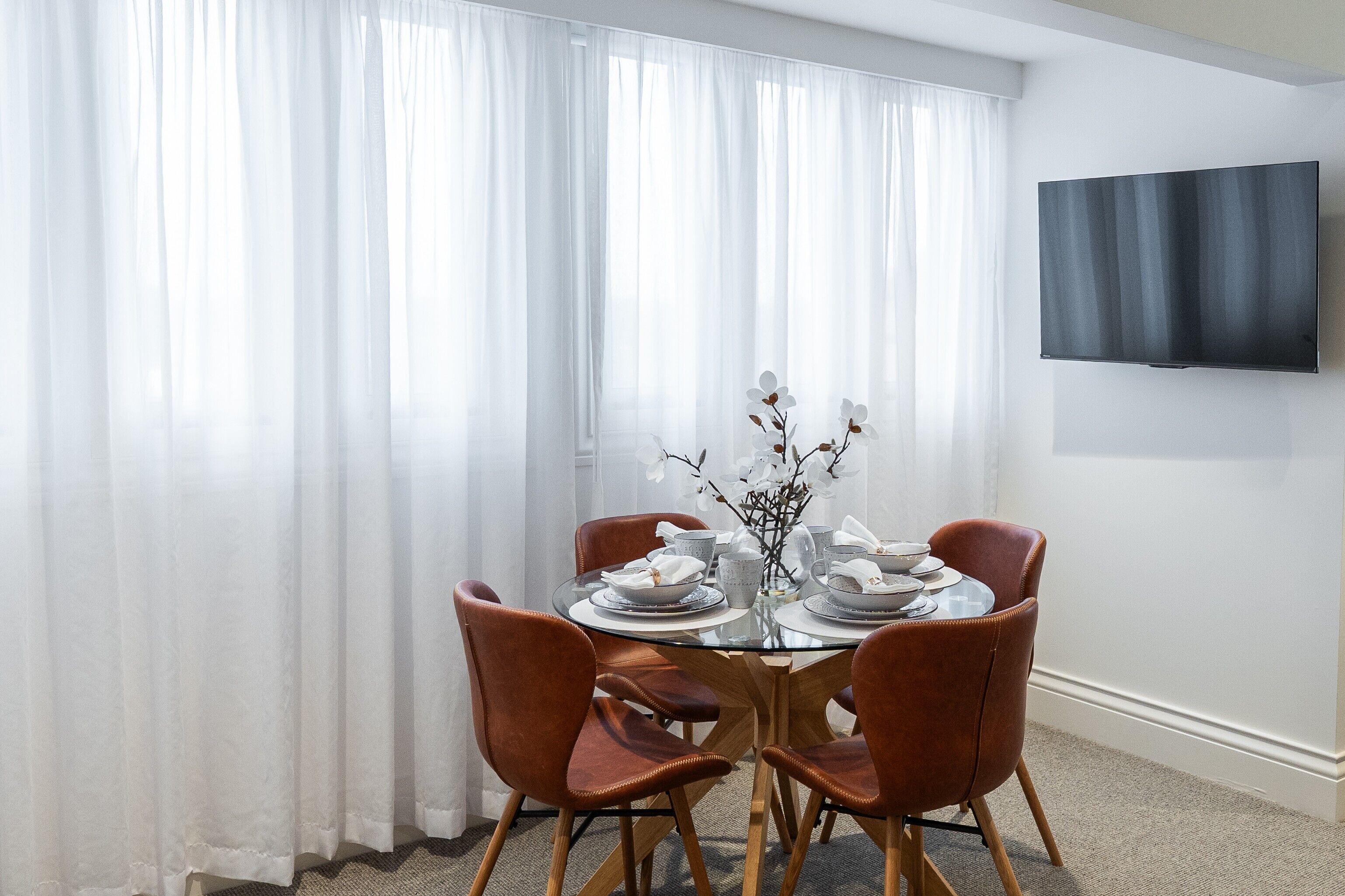 Un coin repas avec une table en verre, quatre chaises marron, des rideaux blancs et un téléviseur à écran plat fixé au mur.