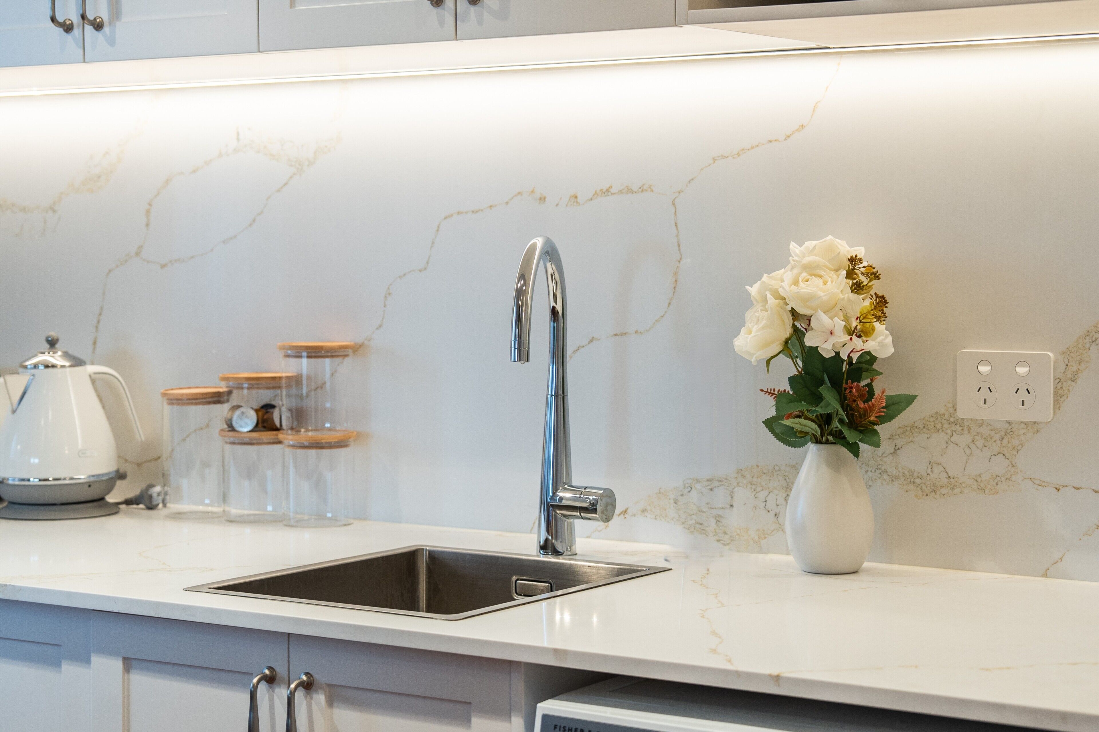 Uma cozinha com pia de aço inoxidável, backsplash de mármore branco e um vaso com flores sobre o balcão.