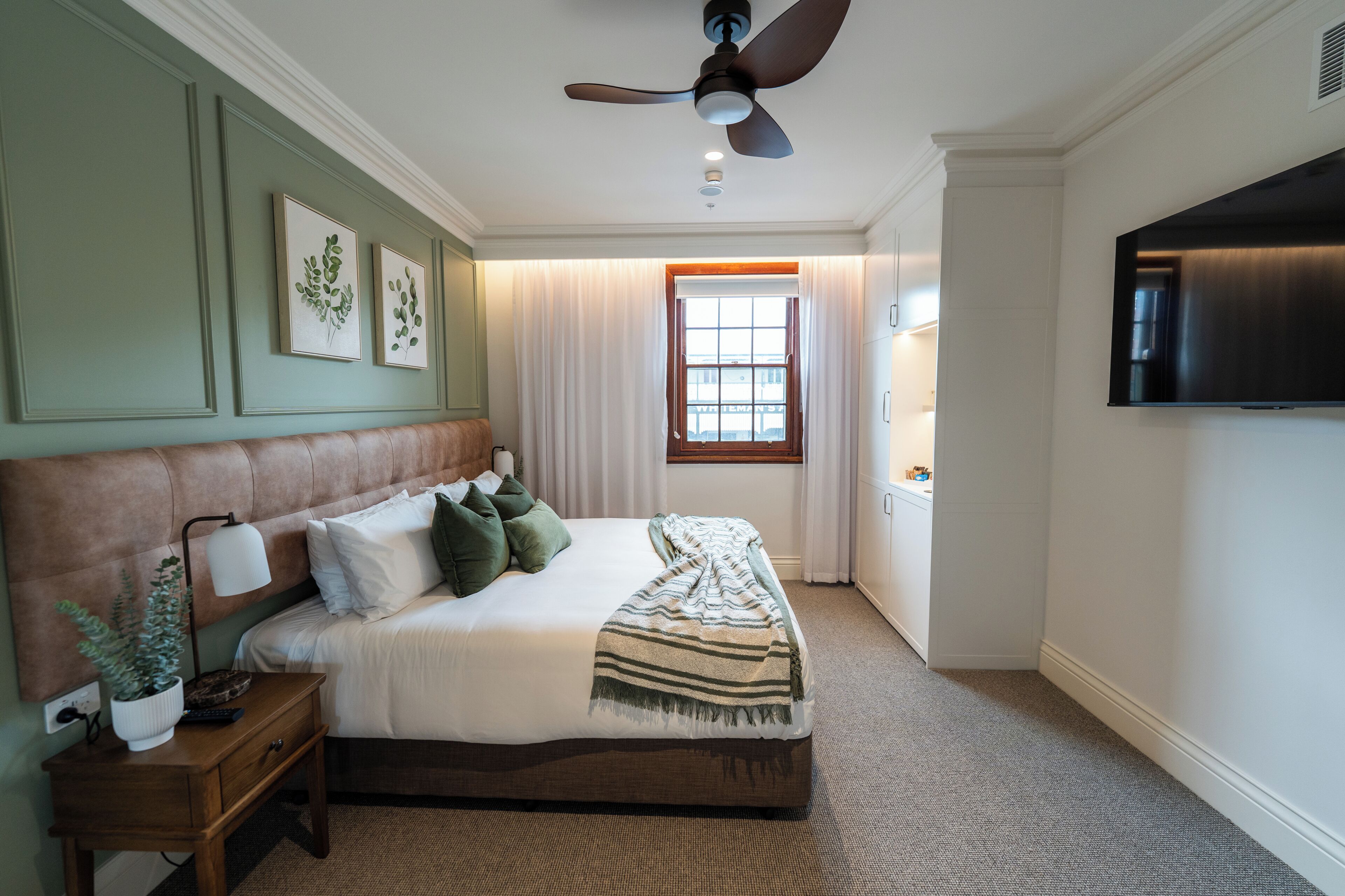 Ein Schlafzimmer mit Bett, Nachttisch, Lampe, Deckenventilator, Fenster und Fernseher.