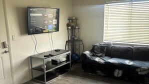Uma sala de estar com uma TV de tela plana na parede, um sofá preto com padrão de folhagem e uma pequena mesa com uma planta.
