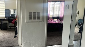 Quarto com piso de madeira, cama com colcha rosa, escrivaninha com computador e uma cadeira.
