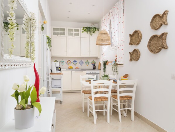 A modern kitchen with white cabinets, a dining area with white chairs, and a small table. - Citrus Garden House (Piano di Sorrento)