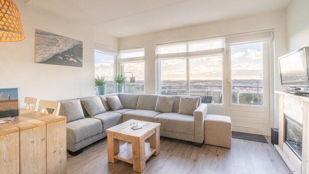Ein modernes Wohnzimmer mit Ecksofa, Couchtisch aus Holz und Fernseher. Große Fenster bieten Blick auf die Stadtlandschaft.