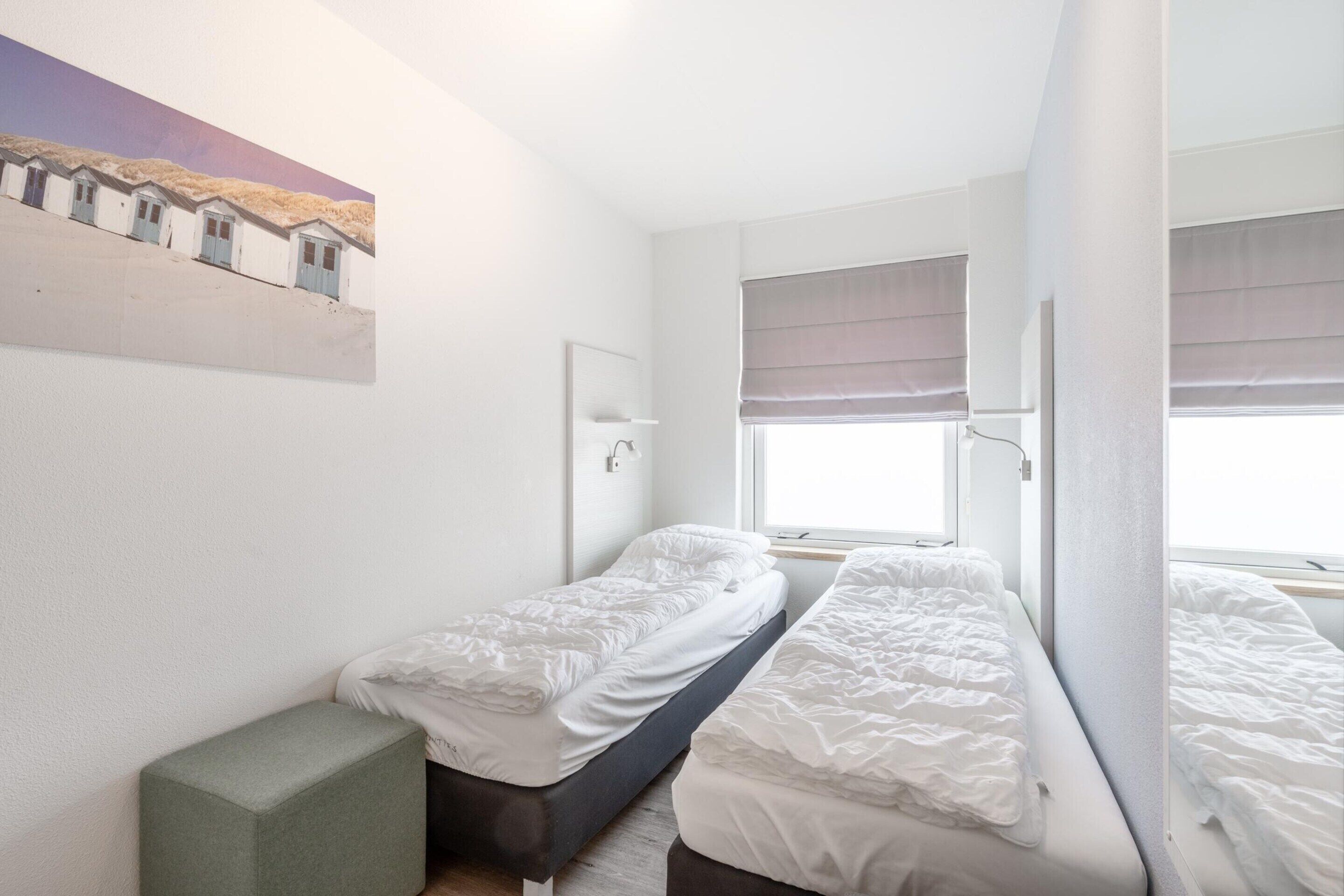 A small, clean room with two single beds, a window with blinds, a green bench, and a painting on the wall.