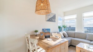 Ein modernes Wohnzimmer mit einem Holzesstisch, weißen Stühlen, einem beigen Sofa und einem großen Fenster mit Blick auf die Stadt.