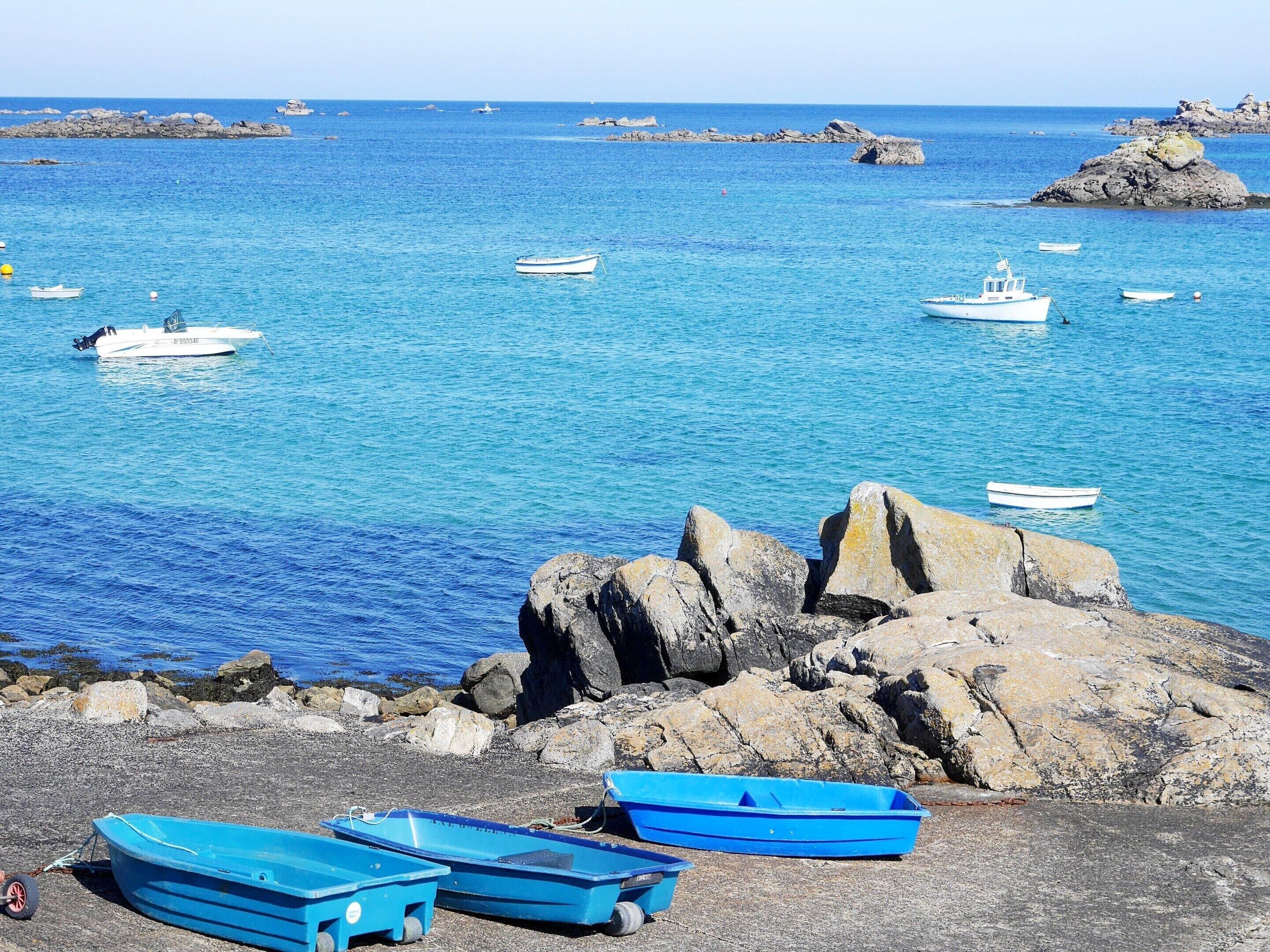 A rocky shoreline with several boats, including a blue rowboat and a white motorboat, near calm blue waters.
