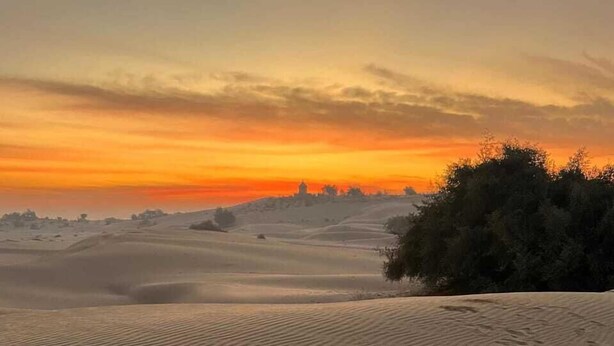 A single bed with a white sheet and pillow in a desert setting with sand dunes and a sunset in the background. - Desert View Resort (Jaisalmer)