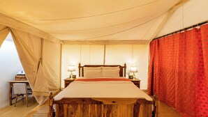 A wooden bed with a beige bedspread, two bedside tables with lamps, a desk with a chair, and a red curtain. - Desert View Resort (Jaisalmer)