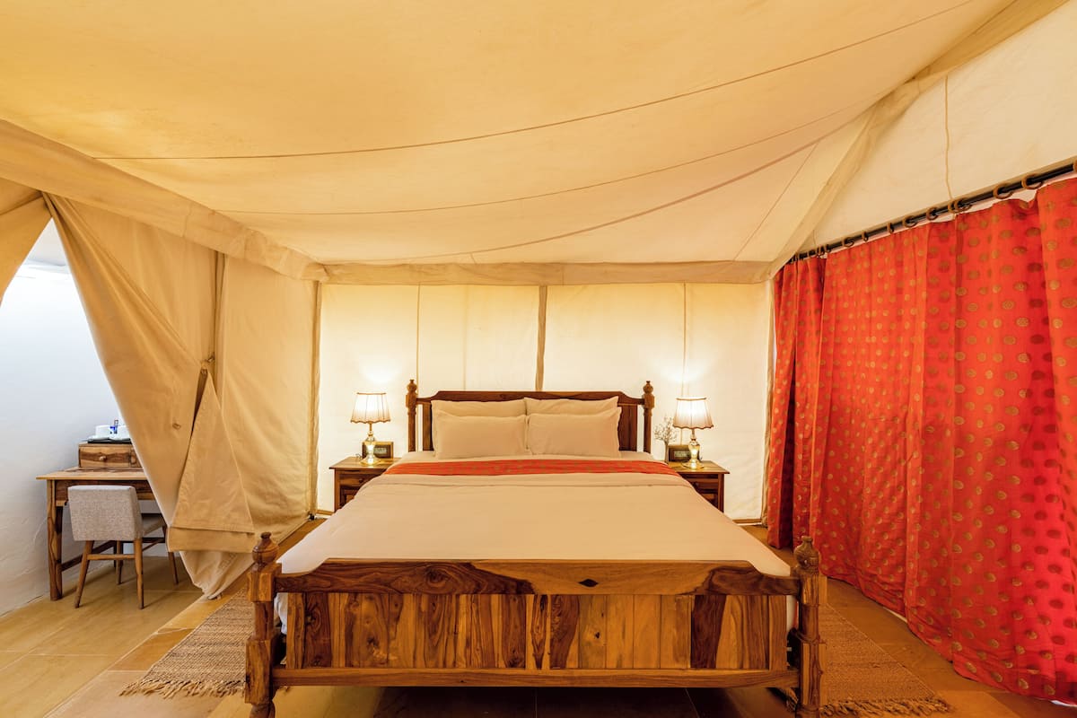 A wooden bed with a beige bedspread, two bedside tables with lamps, a desk with a chair, and a red curtain.