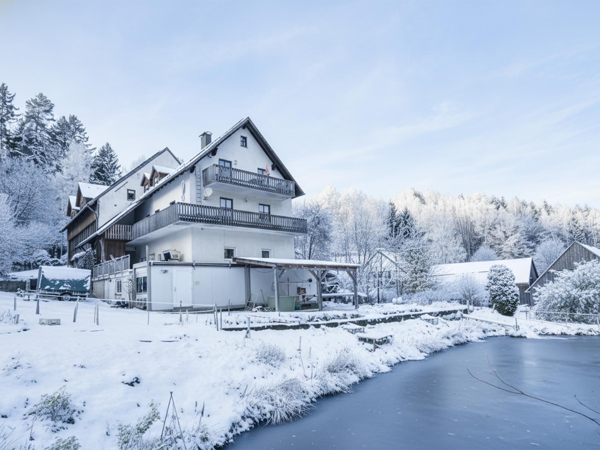 Un chalet à deux étages doté d’un balcon, entouré d’arbres couverts de neige et bordé par une rivière gelée.