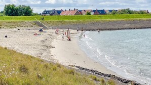 En strand där människor njuter av sanden och vattnet, ett grönområde och hus i bakgrunden.