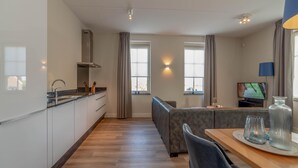 Et moderne køkken med indbygget ovn, et spiseområde med bord og stole, og en opholdsstue med sofa og fjernsyn.