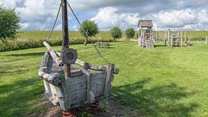 Eine hölzerne Bootskonstruktion mit Seilmechanismus, eingebettet in eine Wiese mit einem Spielhäuschen und einem Picknicktisch im Hintergrund.