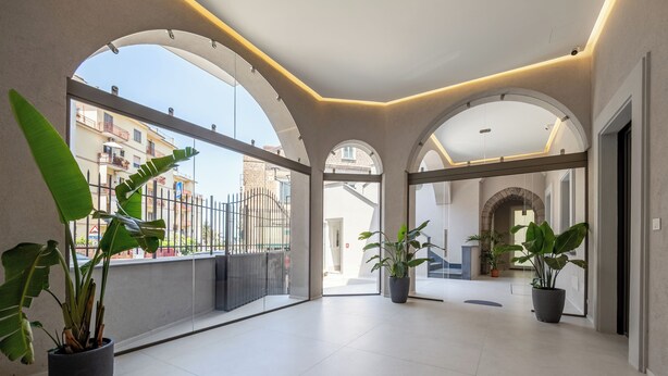 Un pasillo amplio con techos abovedados y ventanas grandes, plantas en macetas y vistas de los edificios exteriores.