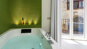 Un baño moderno con una pared verde con textura, una bañera y vistas a un balcón con un edificio rústico.