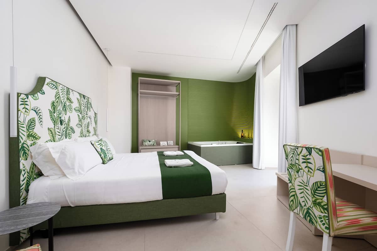 A modern hotel room with a large bed, a desk, a chair, a TV, a bathroom with a bathtub, and a green accent wall.