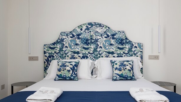 Un lit bien fait, avec une couvre-lit bleue et des oreillers blancs, une tête de lit ornée d’un motif floral et une table de chevet avec une lampe.