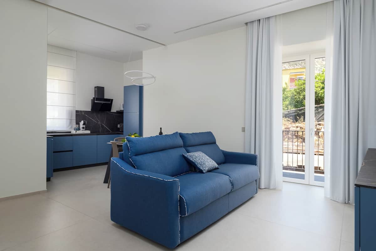A modern living room with a blue sofa, a kitchen area with dark cabinets, and a view of the outside through large windows.