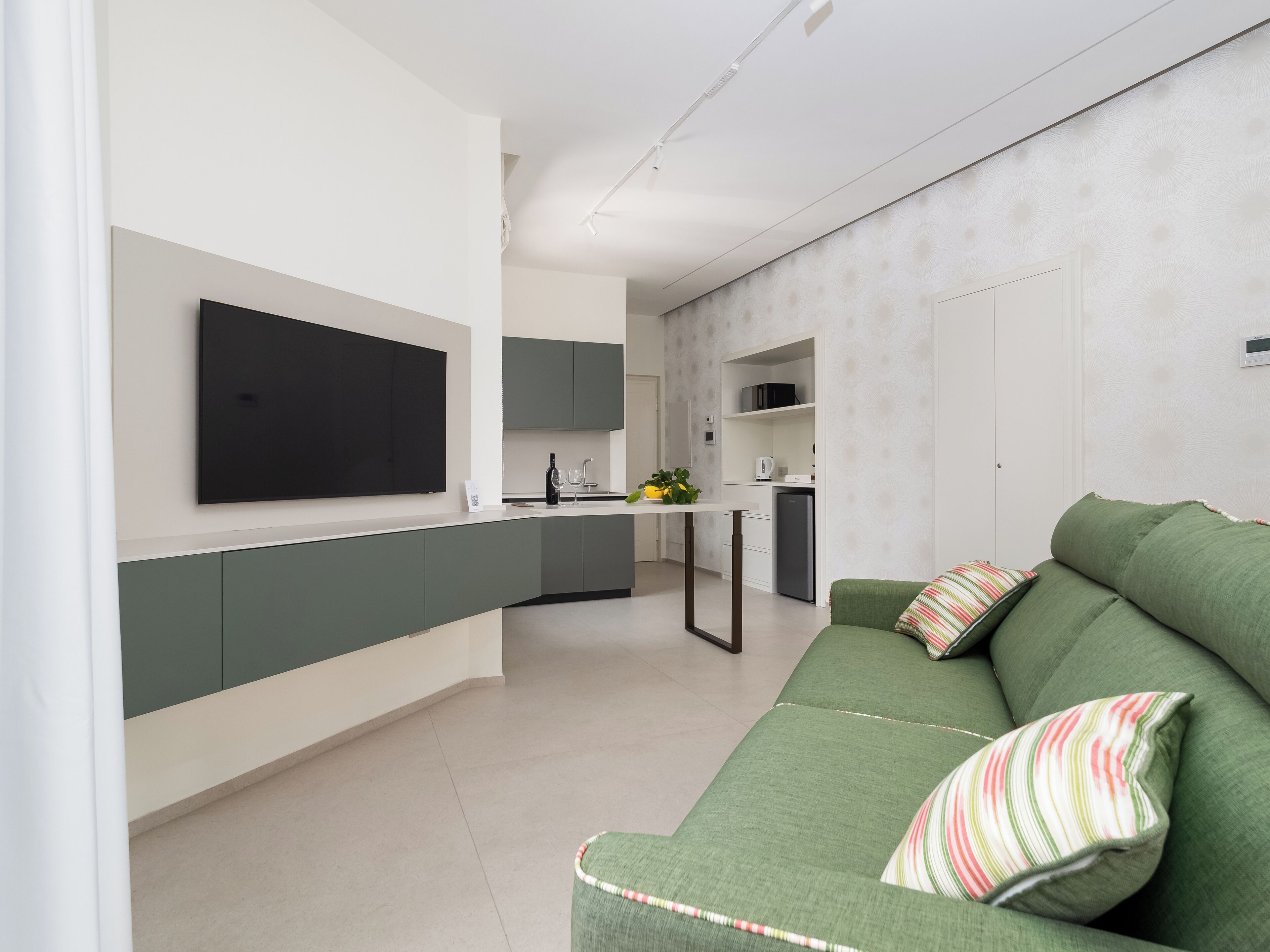 A modern living room with a green sofa, a flat-screen TV mounted on the wall, and a kitchen area with a white countertop and green cabinets.