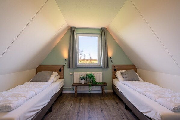Une petite chambre mansardée triangulaire, avec deux lits simples, une fenêtre avec des rideaux, un radiateur et une petite table sur laquelle est posée une plante.
