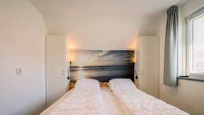 A twin bed with white bedding, a headboard with a sunset painting, and a window with curtains. - Tastefully and Modernly Furnished Holiday Home for 6 Persons (De Waal)
