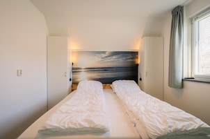 Uma cama de casal com roupa de cama branca, cabeceira com pintura de pôr do sol e janela com cortinas.