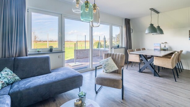 A modern living room with a sofa, a dining table with chairs, and a view through large windows. - Tastefully and Modernly Furnished Holiday Home for 6 Persons (De Waal)
