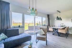 Uma sala de estar moderna com sofá, mesa de jantar e cadeiras, e vista através de grandes janelas.