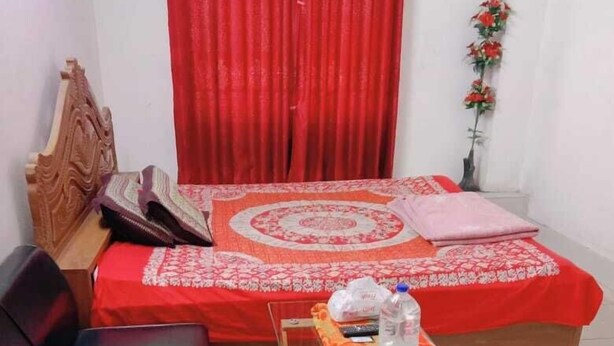 Una habitación pequeña y bien iluminada con una cama, un sofá negro, una mesa de vidrio con una botella y una cortina roja.