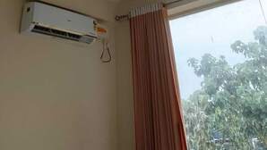 A room with a ceiling fan, an air conditioning unit, a window with a curtain, and a view of trees outside. - SOHAN BOARDING AND LODGING (Udupi)