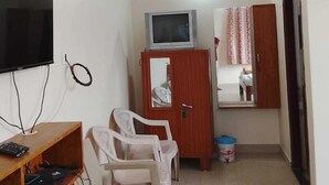 A small, sparsely furnished room with a TV, a wooden table, a white chair, a cabinet with a mirror, and a small rug on the floor. - SOHAN BOARDING AND LODGING (Udupi)