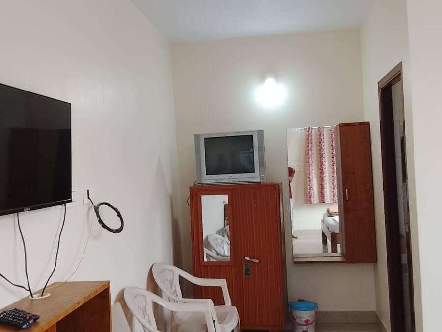 Una habitación pequeña y poco amoblada, con una televisión, una mesa de madera, una silla blanca, un armario con un espejo y una alfombra pequeña en el suelo.