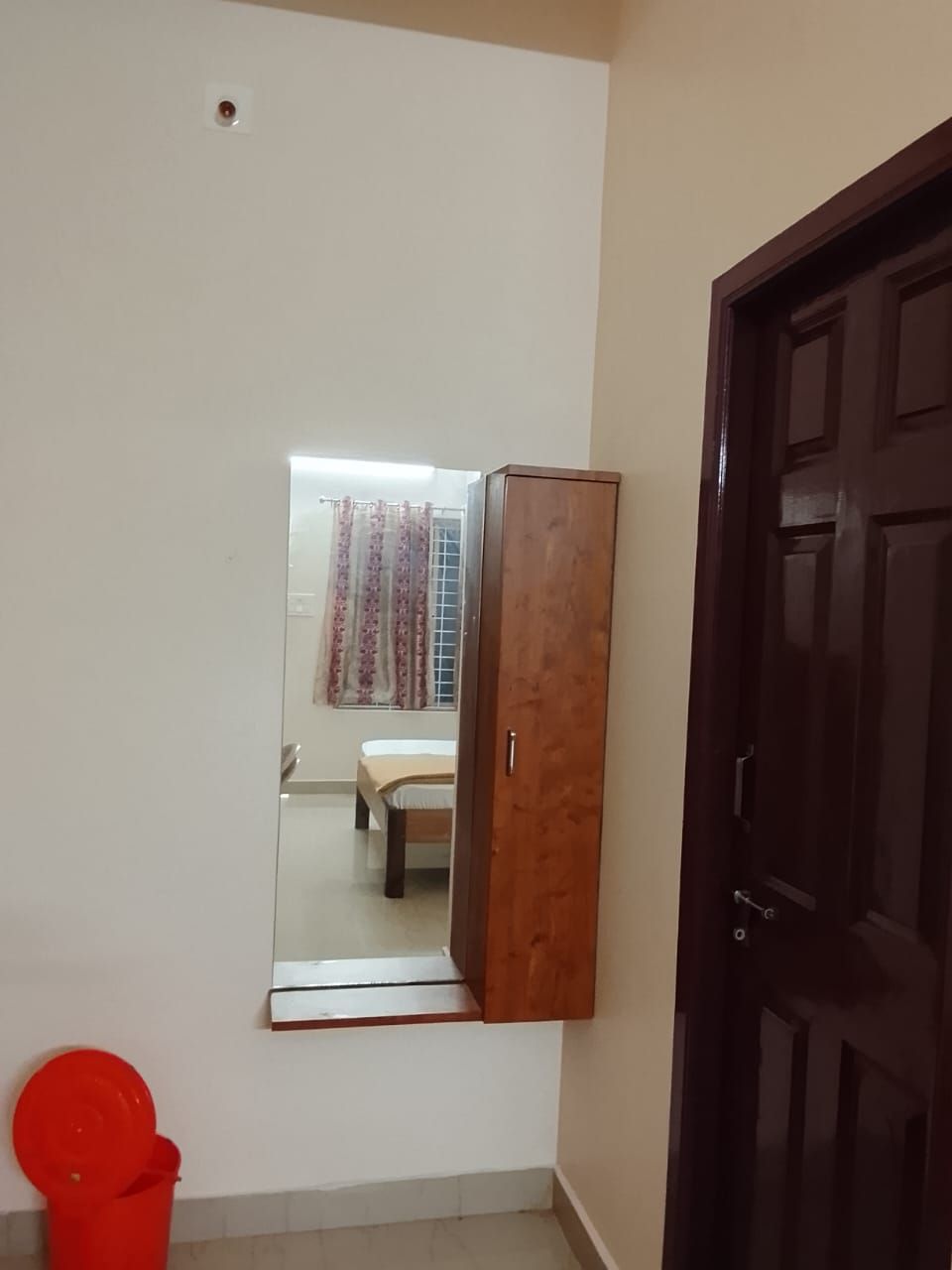 Une pièce avec une armoire en bois, un seau rouge et un miroir qui reflète un lit et des rideaux.