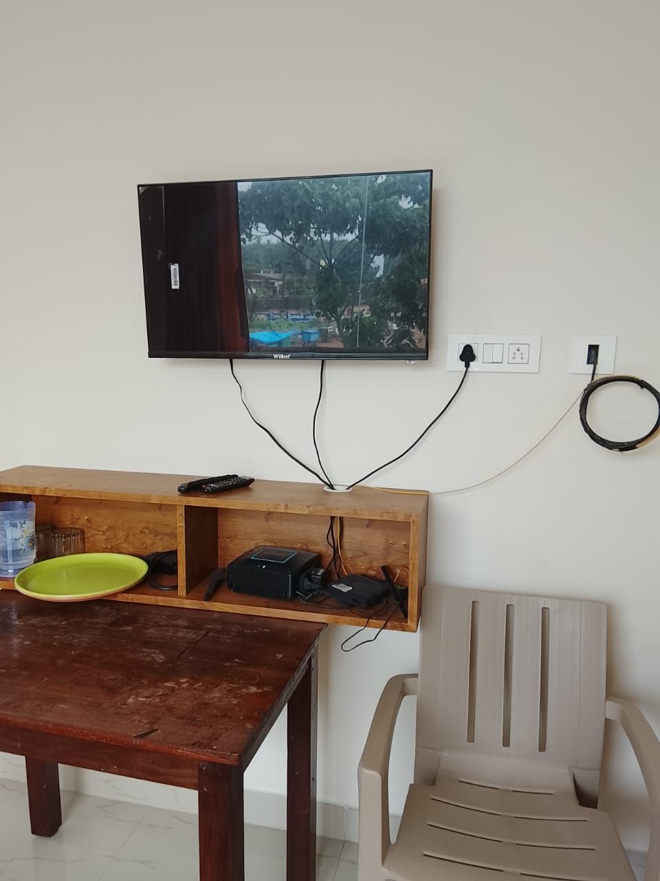 A room with a television mounted on the wall, a wooden table with a green plate, a plastic chair, and a power strip with cables.