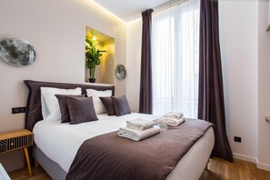 Una cama bien hecha con ropa de cama blanca y marrón, una mesita de noche con lámpara, y un ventanal con cortinas.