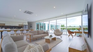 Un salon moderne avec un canapé, des fauteuils et une table basse. Il y a également un coin cuisine avec des meubles blancs et une table à manger. De grandes fenêtres offrent une vue sur l’extérieur.