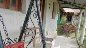 Un espacio al aire libre cubierto con un columpio, suelo de concreto y un pequeño jardín con una silla y una estructura de paja.